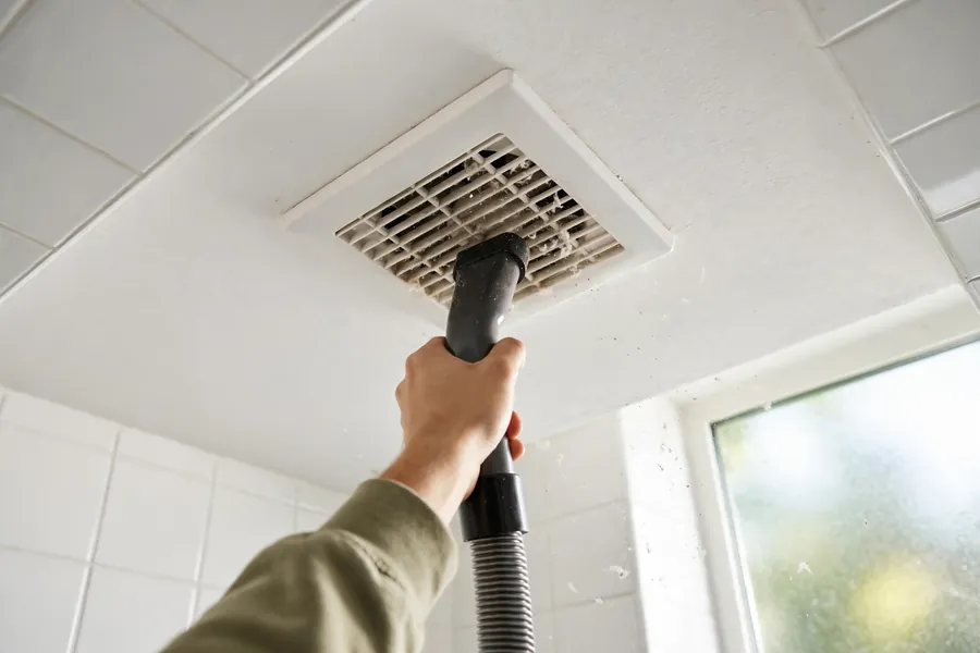Hand vacuuming a white ceiling exhaust fan grille