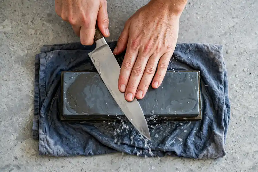Hands pushing a chef's knife forward across a wet whetstone on a damp towel.