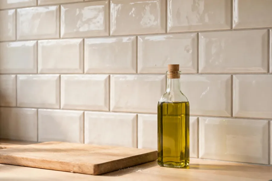 Warm white glazed subway tile backsplash in a running bond above a wood countertop with a cutting board and olive oil bottle